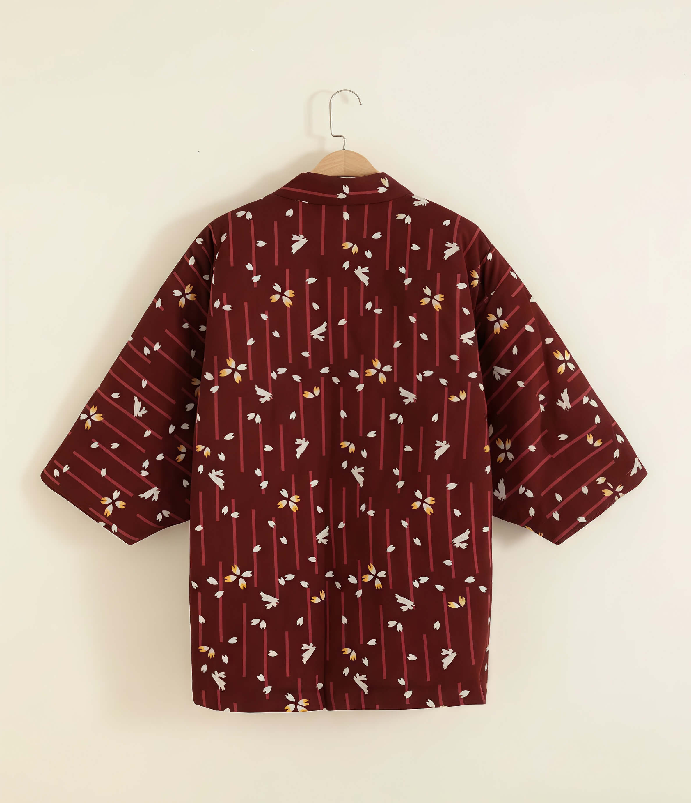 Back view of women’s red Japanese hanten jacket with sakura floral pattern hanging on wooden hanger against neutral warm background